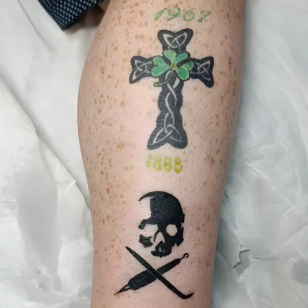Cruz celta con trébol verde y, debajo, una calavera sobre una plumilla y un gancho cruzados, en la pierna